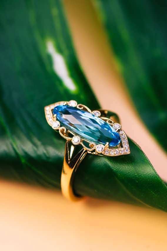 Marquise blue topaz ring in ornate gold setting with natural diamond scrollwork, on a green leaf
